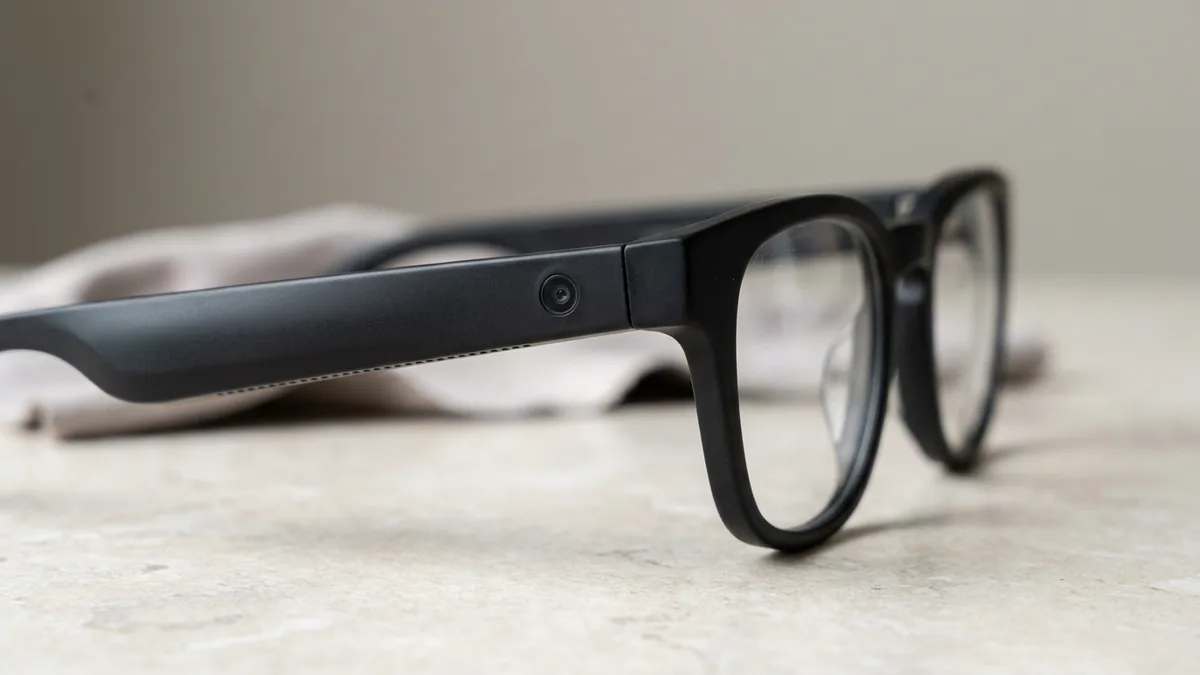 Close-up of matte black smart glasses showing the tiny camera lens and speaker holes built into the temple arm, with plain clear lenses and no display