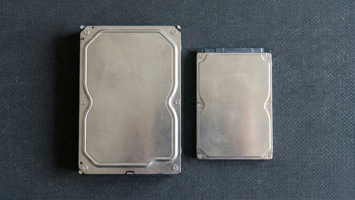 A 3.5-inch desktop hard disk and a smaller 2.5-inch laptop hard disk lying side by side on a dark textured antistatic mat, both with their metal lids closed and completely unmarked, seen from directly above.