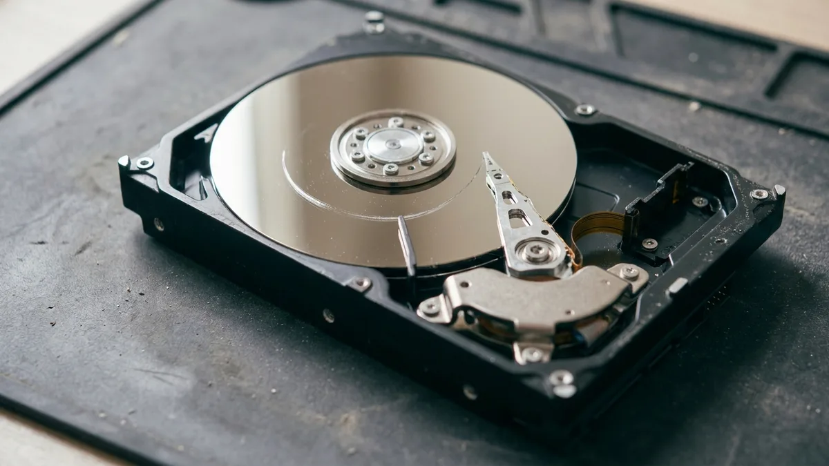 An opened 3.5-inch hard disk on a dark mat with its lid removed, showing the mirror-finish platter, the central spindle hub and the head arm swung out across the disc, with a curved scratch scored into the platter surface.
