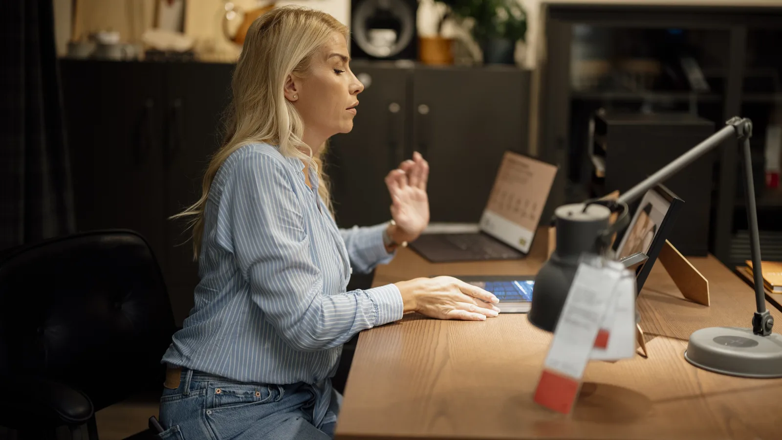 A blonde woman in a light-blue striped shirt sits at a wooden home-office desk, one hand raised as she works, with an open laptop, a desk lamp and a small tablet stand around her.