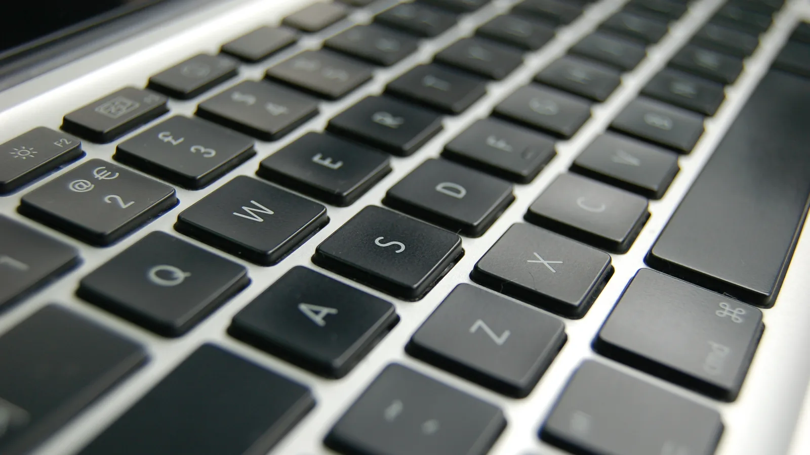 An extreme close-up of a laptop keyboard with black keys on a silver body, the Q, W, E, A and S keys in sharp focus and the rest of the rows falling out of focus.
