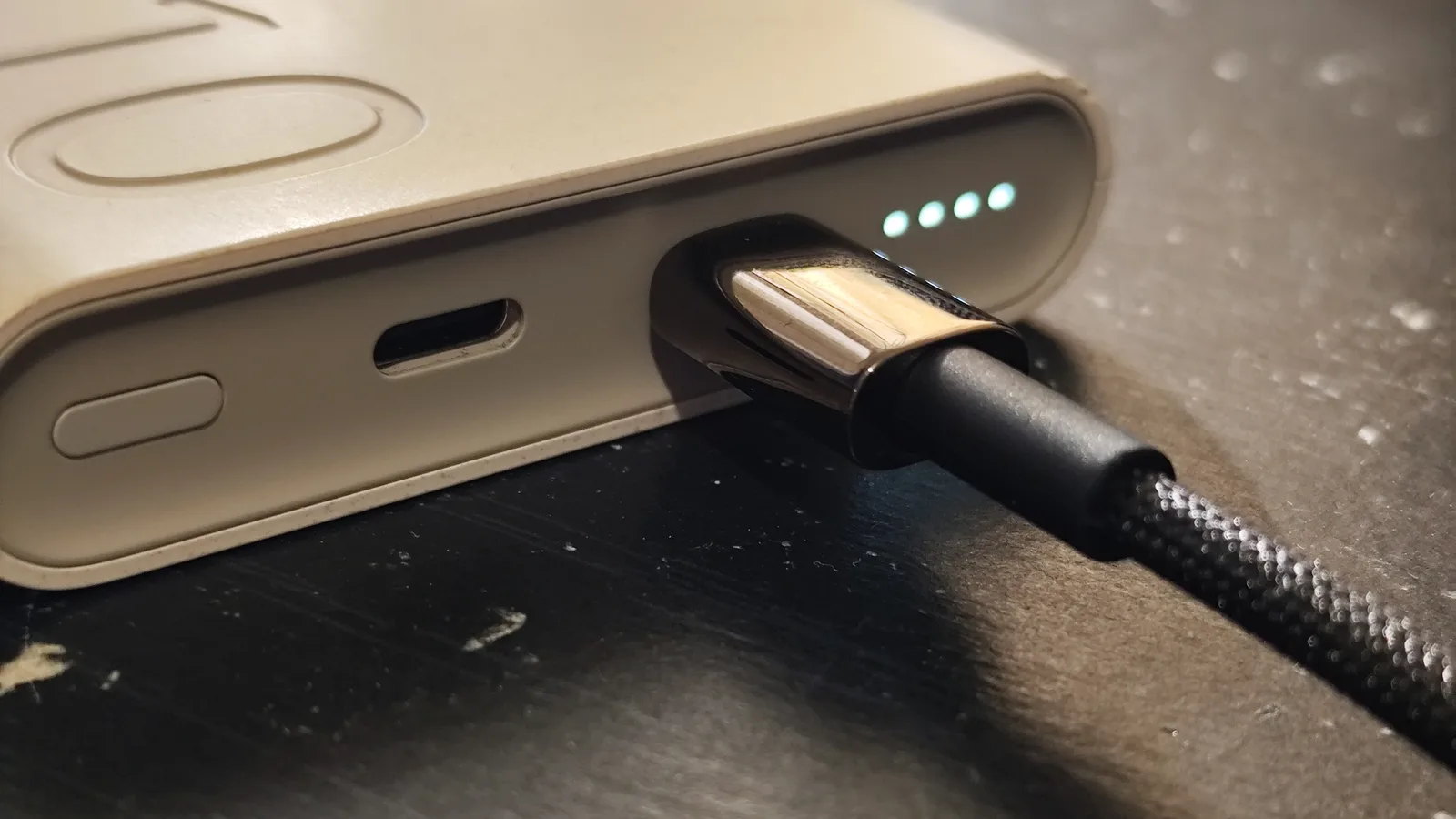 A black braided charging cable plugged into a beige power bank, next to an empty USB-C port and a button, with four small charge-level lights lit