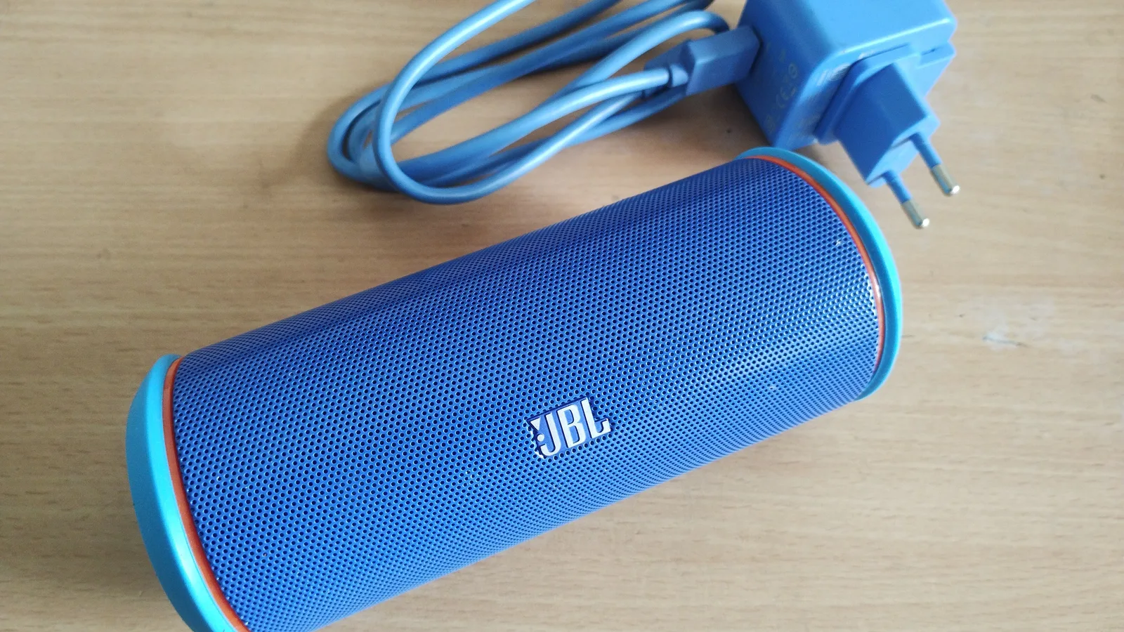 A blue JBL portable speaker lying on a wooden table beside its coiled blue charging cable and a blue wall adapter with a two-pin plug