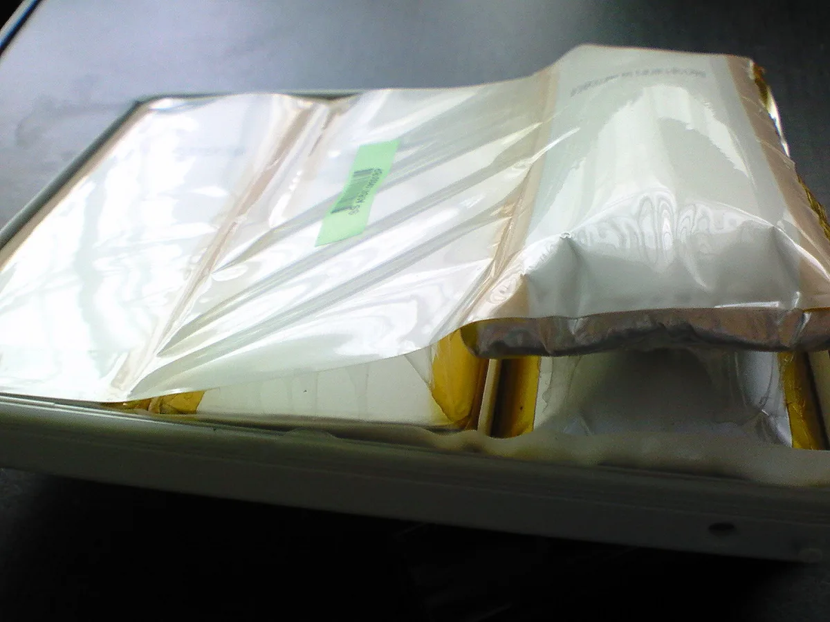 Pale swollen pouch cells bulging up out of a laptop battery pack's plastic housing, with gold-coloured foil edges and a small pale-green label between them