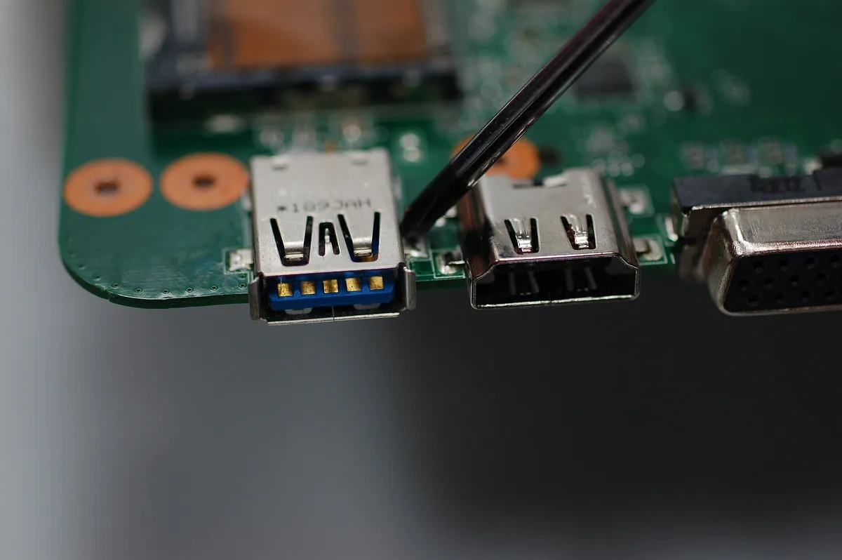 A metal probe touching the edge of a dark green laptop motherboard beside a row of soldered sockets, with copper-coloured pads on the board nearby