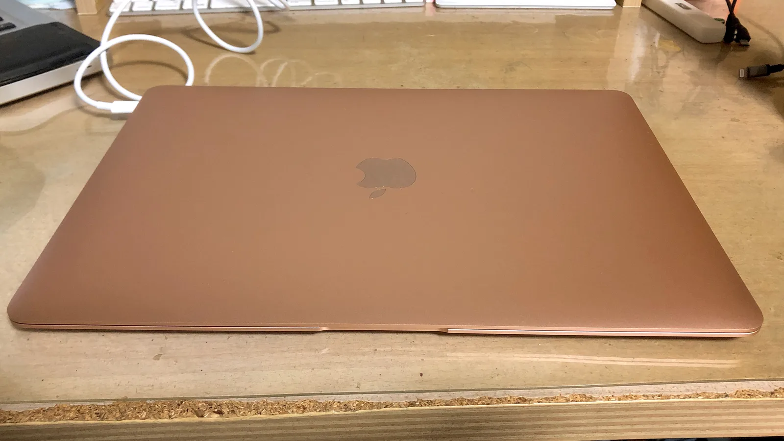 A closed gold Apple laptop lying on a wooden desk, with a white charging cable plugged into its left side