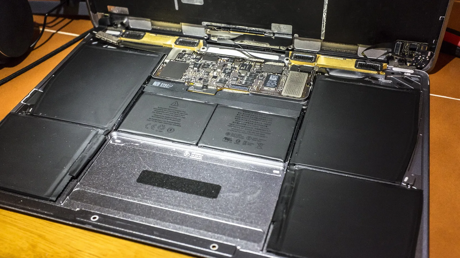 Inside a space-grey Apple laptop with its bottom case off: six flat black battery cells fill most of the body around the metal back of the trackpad, with a small main circuit board just below the hinge