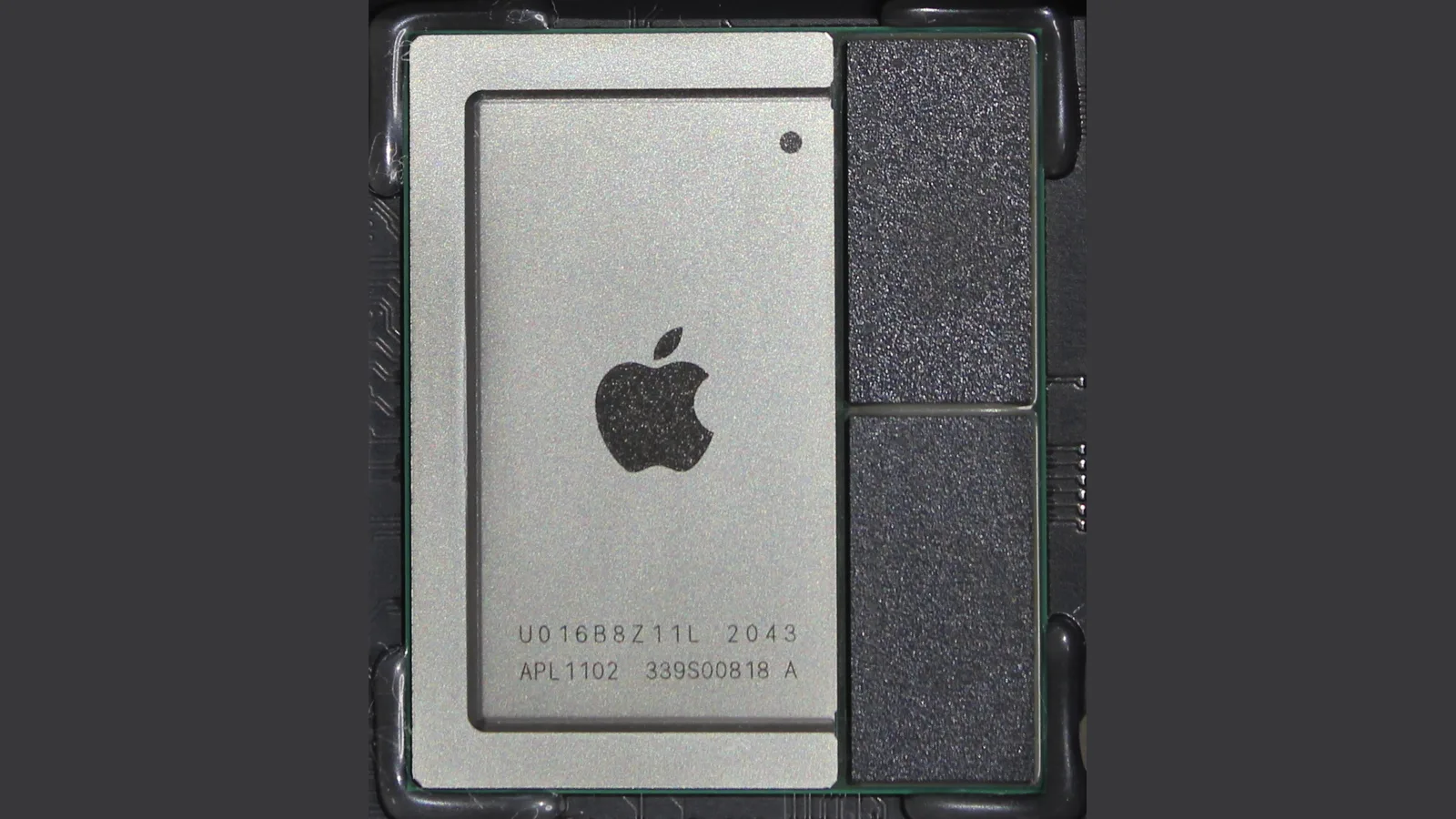Gambar dekat sebuah cip Apple pada papan litar gelap: penutup perak dengan logo Apple dan tanda APL1102, serta dua cip memori hitam di sebelahnya