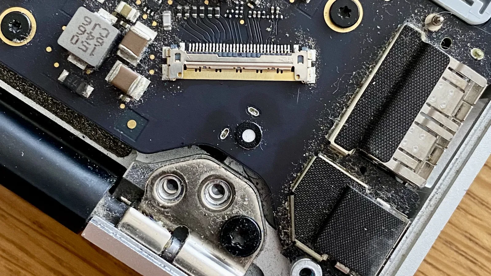 Bahagian dalam komputer riba Apple yang dibuka, di sebelah engsel skrin: soket kosong pada papan litar gelap tempat kabel dipasang, titik putih kecil dalam gelang hitam di bawahnya, dan habuk di seluruh papan.