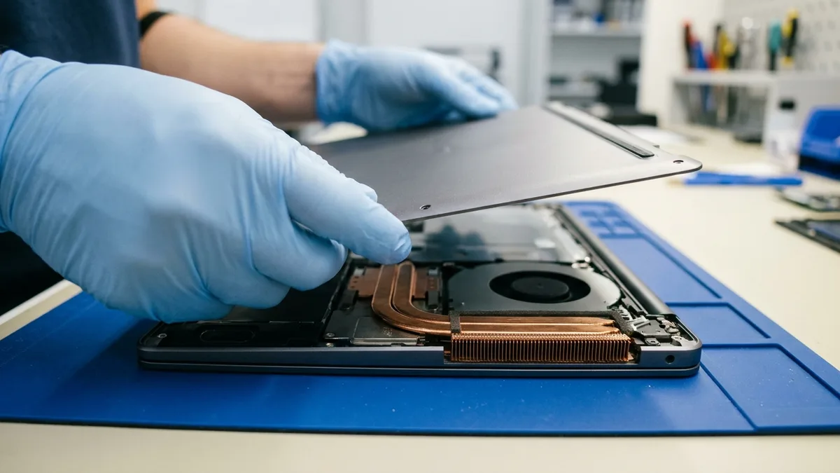 Hands in pale blue gloves lifting the plain bottom cover clear of a slim dark grey laptop on a deep blue anti-static mat, uncovering the copper heat pipes, a stack of fine copper cooling fins and a black fan inside.