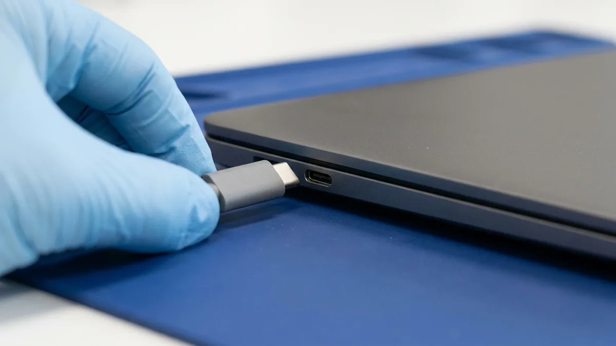 A hand in a pale blue glove guiding a plain grey USB-C connector towards the port on the side edge of a slim closed dark laptop resting on a deep blue anti-static mat.