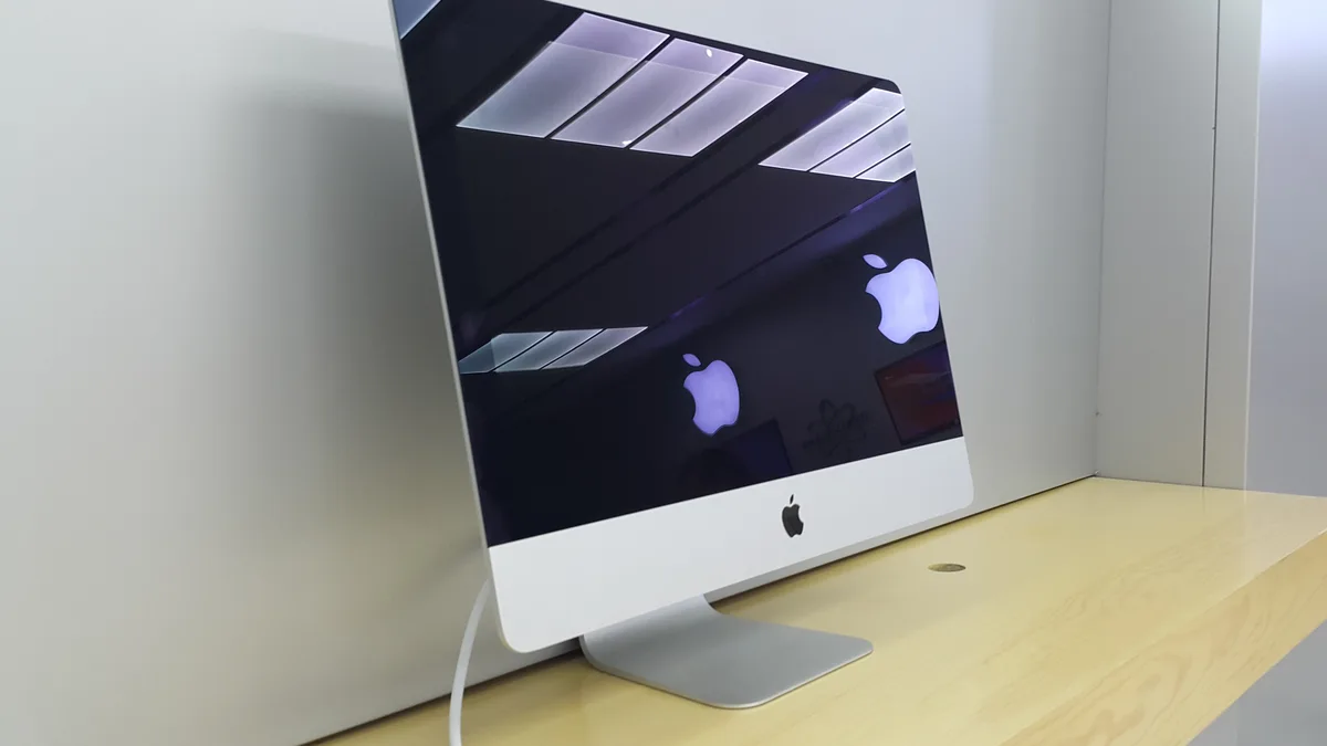 A slim aluminium iMac standing on a pale wooden shelf with its display completely dark, the ceiling lights reflecting off the black glass