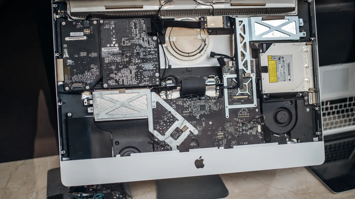 An aluminium iMac opened at a repair service with its display panel lifted away, showing the logic board, speakers, cooling fans and optical drive behind it