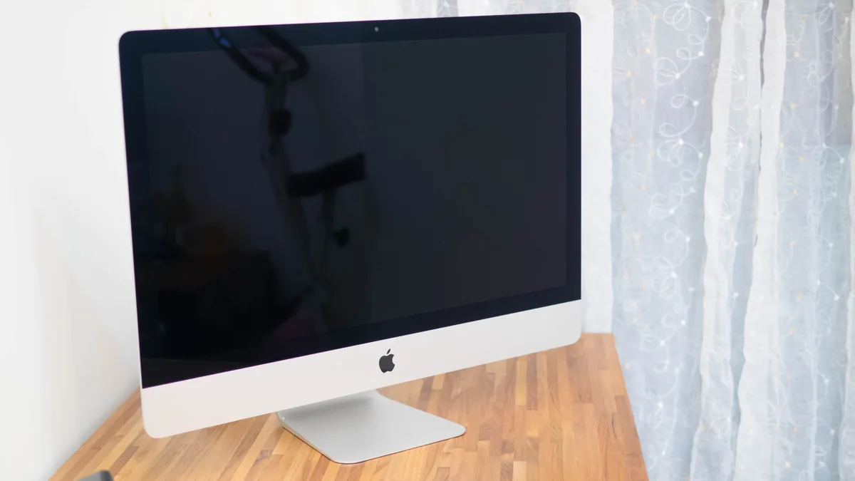 A 27-inch aluminium iMac standing on a clean wooden desk beside a curtained window, its screen dark and unmarked