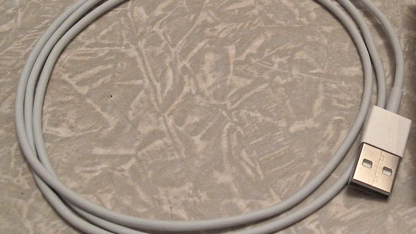 Kabel pengecas putih digulung menjadi bulatan, dengan palam USB-A logam leper di latar depan pada permukaan kelabu berbelang