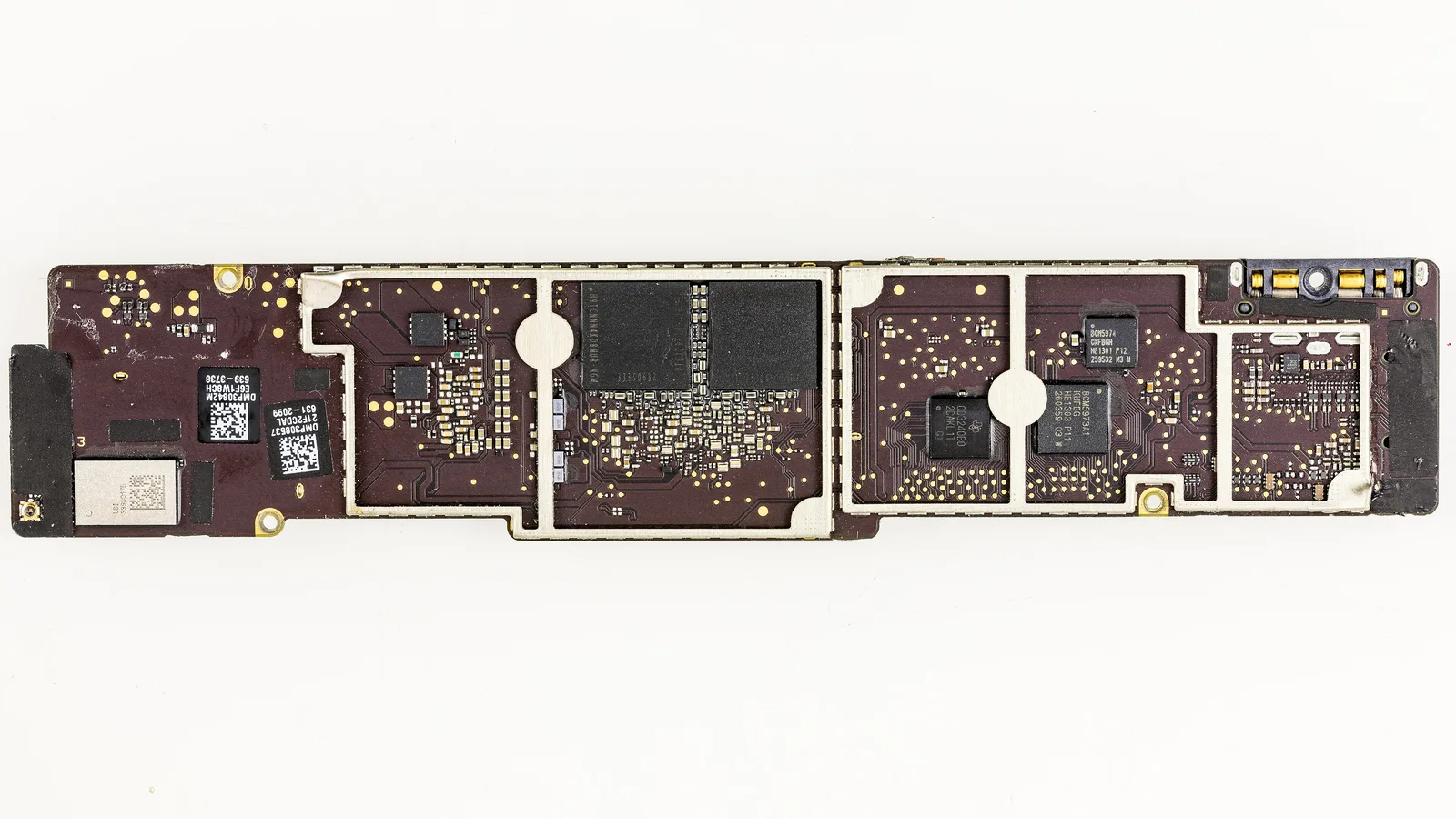 The bare back of a long, narrow logic board on a white background: a dark maroon circuit board divided by silver metal shields, two large black chips in the middle, smaller black square chips on the right, two black barcode labels and a silver module on the left, and a connector with gold contacts at the top right