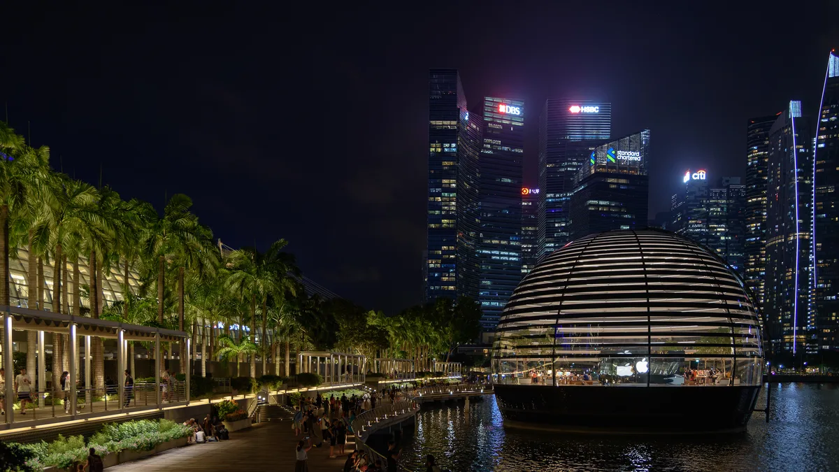 Kubah kaca dan keluli yang bercahaya dari dalam, berdiri di atas air gelap di sebelah promenad tepi air yang bercahaya, dengan menara-menara ufuk bandar di belakangnya pada waktu malam