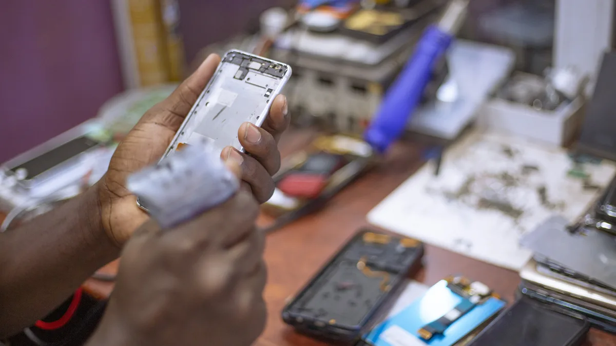 Sepasang tangan memegang rangka logam telefon mudah alih yang telah dibuka di atas meja kerja kayu, dengan alat pengumpil plastik biru dan beberapa telefon yang telah dileraikan disusun di sebelahnya