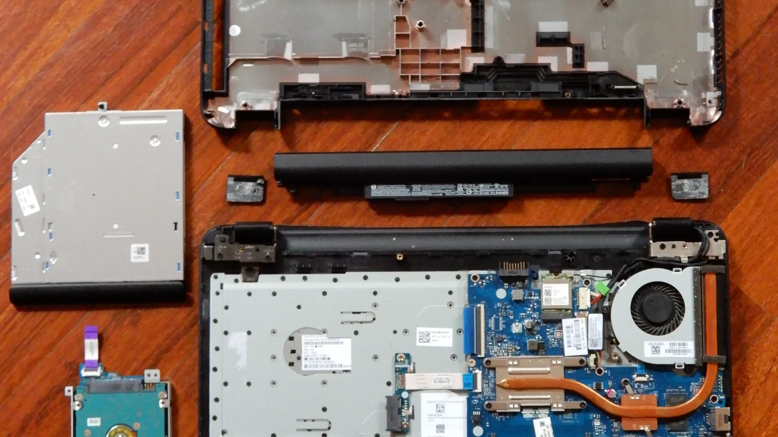 A laptop taken fully apart on a wood-grain surface, its parts laid out separately — the bottom cover, a black battery, an optical drive, a hard drive, and the main chassis with a blue motherboard, copper heat pipe and cooling fan