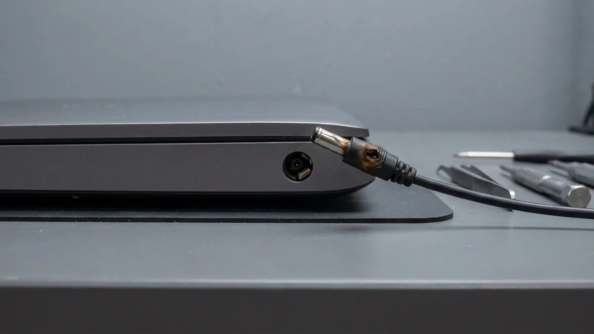 A close-up of a laptop's round charging socket with the charger's barrel plug held just short of it: the plug's metal tube is bent to one side and its collar is scorched brown.