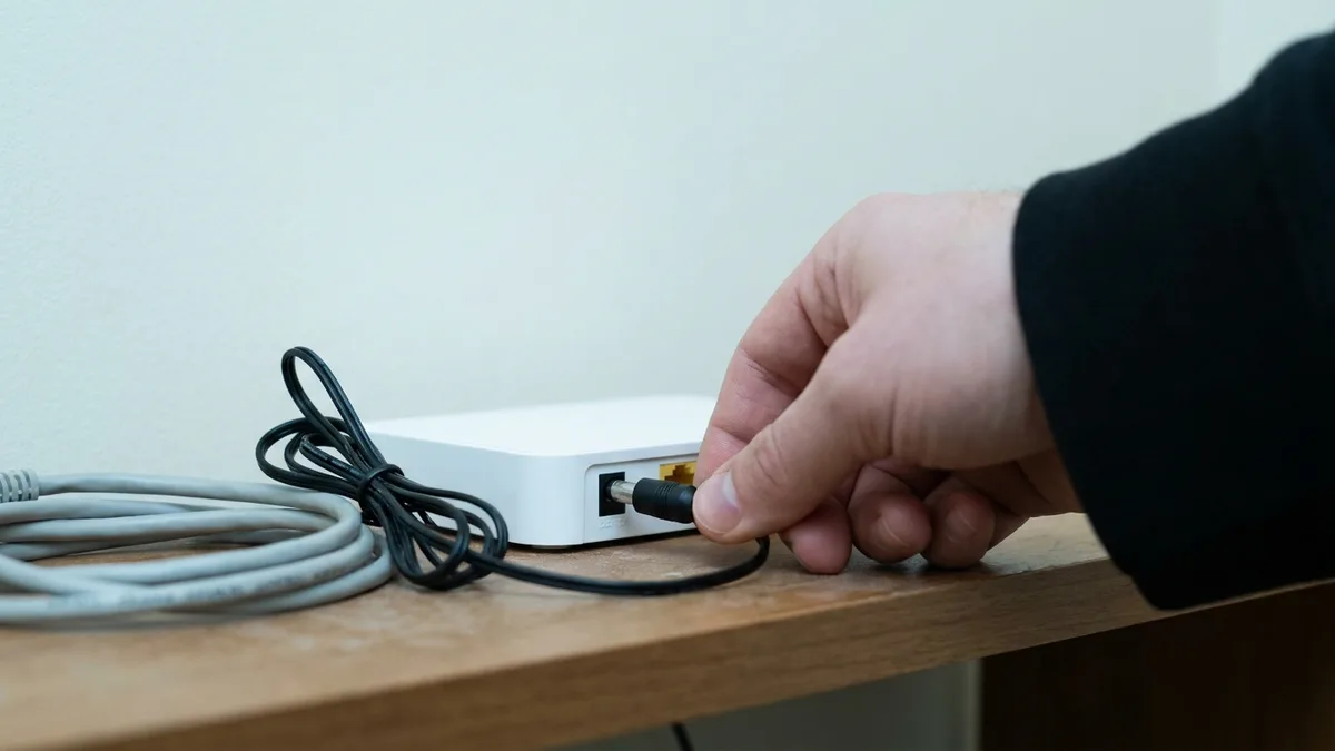 A hand pulls the black power plug out of the back of a small plain white router standing on a dusty wooden shelf, the plug half withdrawn from its socket, with a coiled pale network cable and the slack black power cable lying beside it