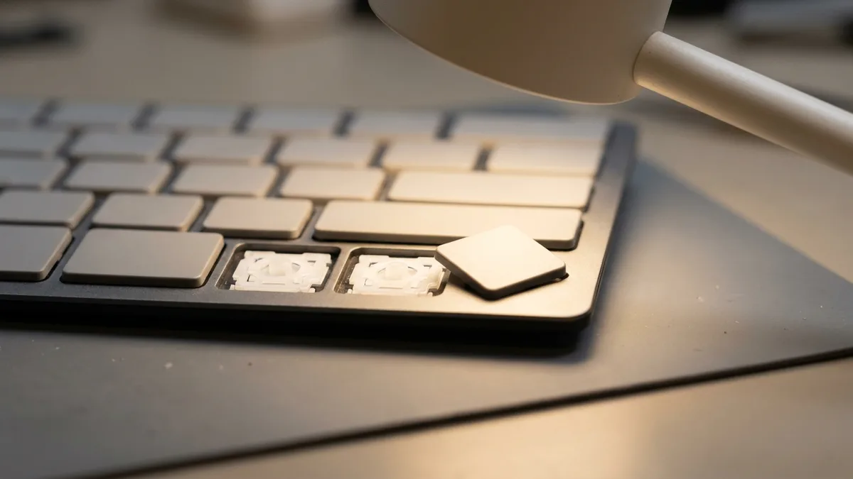 Gambar dekat satu penjuru papan kekunci: dua penutup kekunci hilang sepenuhnya sehingga mekanisme gunting putih terdedah dalam petaknya, dan penutup ketiga terlepas sepenuhnya lalu terletak rata pada rangka di sebelahnya.