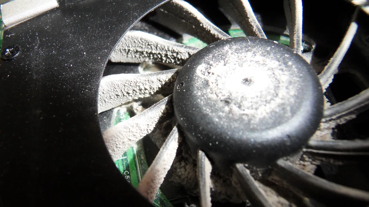 A laptop cooling fan choked with felted grey dust, the blades and the board behind them completely coated