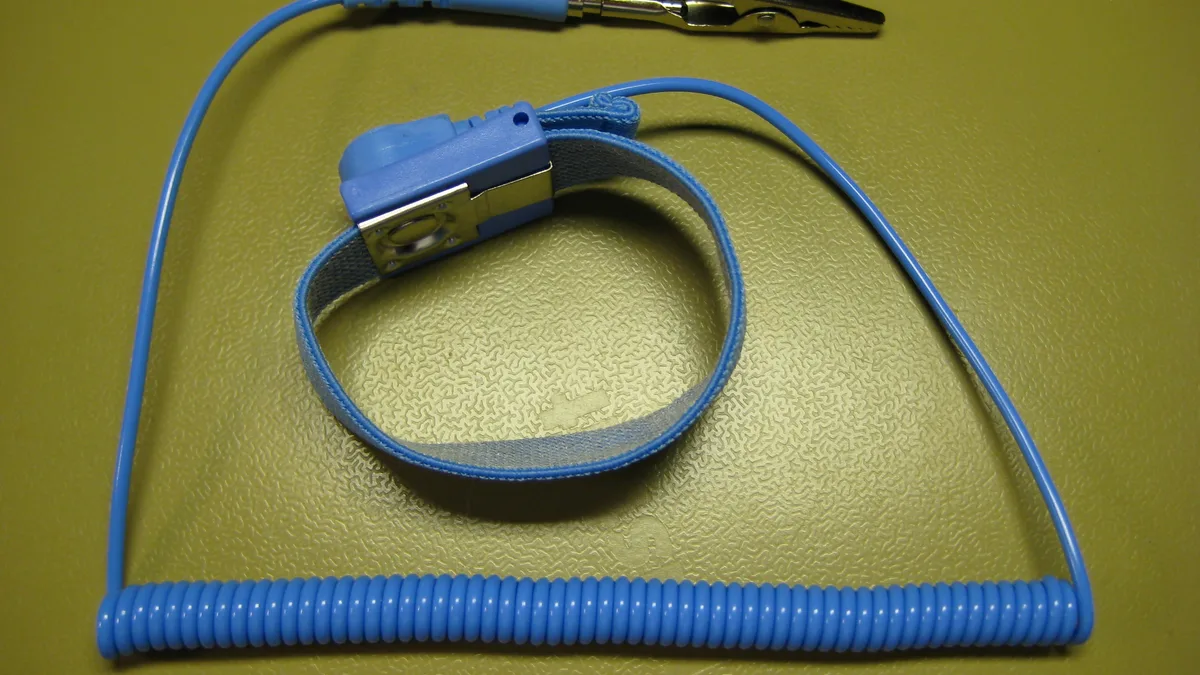 An anti-static wrist strap with its blue elastic band, metal contact plate and coiled lead ending in a crocodile clip