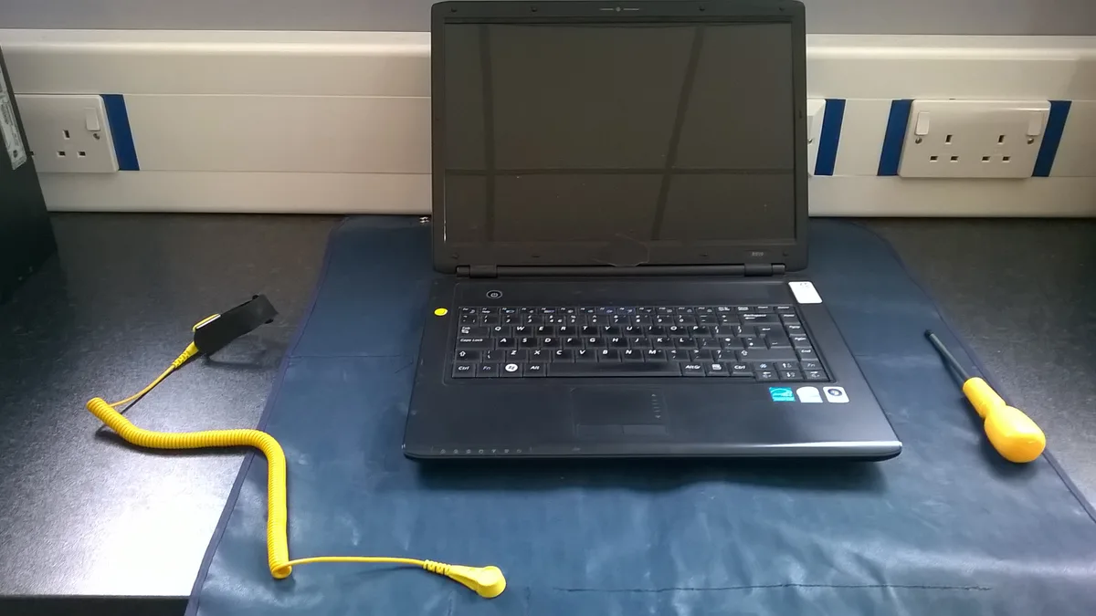 A workbench set up for laptop maintenance: the laptop sits with its screen dark on an anti-static mat, a wrist strap and a screwdriver beside it