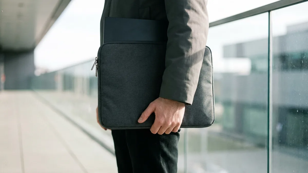A closed slim laptop zipped into a plain dark padded sleeve, carried at the side in one hand along a bright covered walkway beside a plain glass balustrade, the walkway empty and the daylight beyond it thrown out of focus.