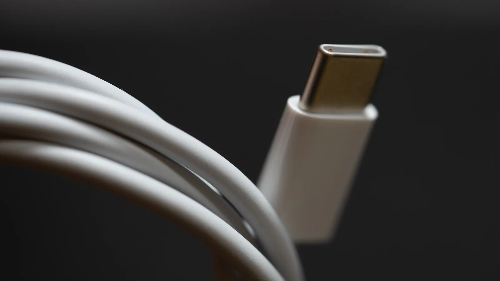 Gambar dekat kabel USB-C putih yang digulung di hadapan latar gelap, dengan palam USB-C bujur leper dan cengkerang logam berwarna emas dalam fokus tajam di sebelah kanan