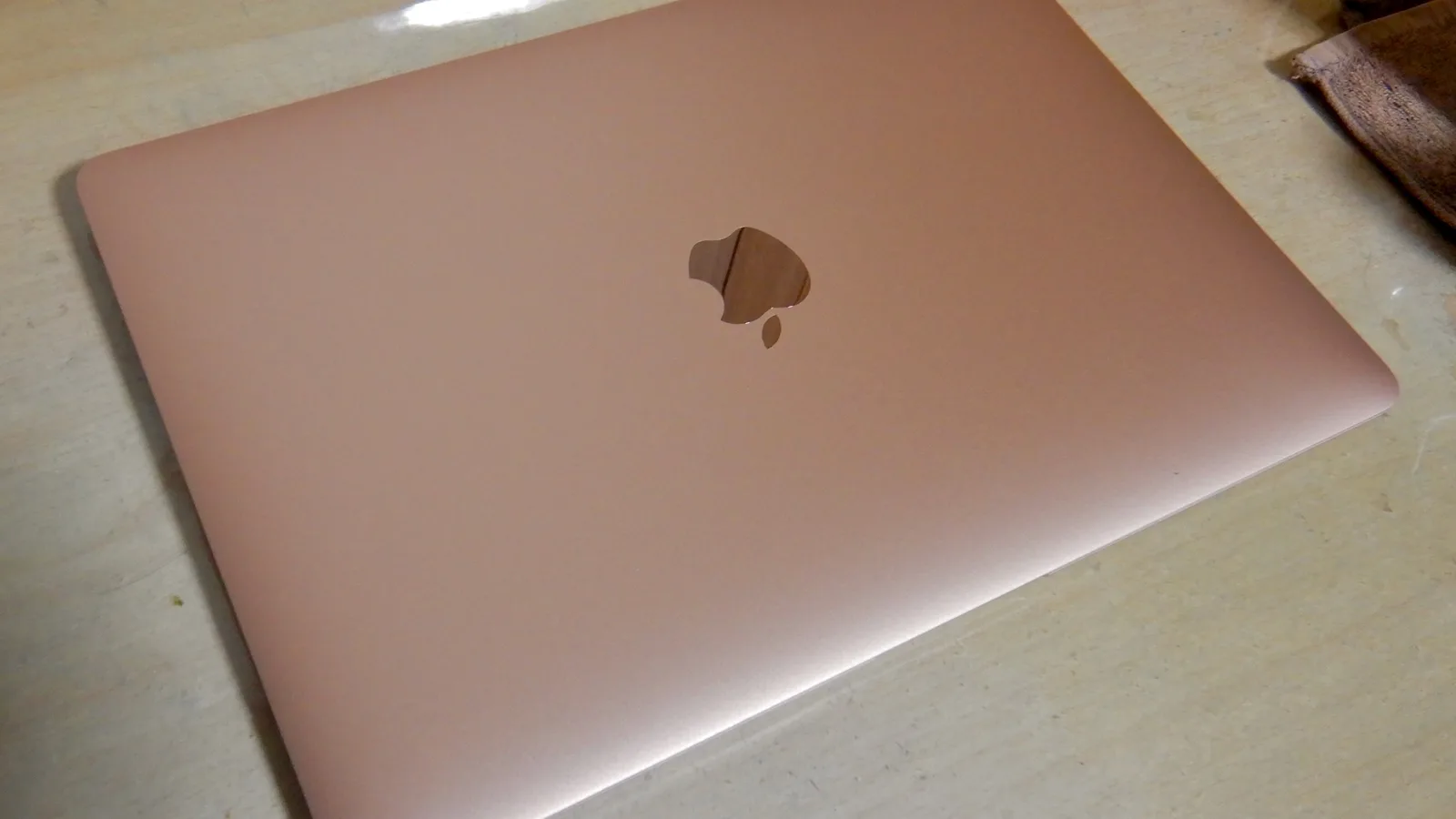 Penutup komputer riba Apple berwarna emas yang tertutup, terletak secara serong di atas meja berwarna cerah, dengan logo Apple berkilat di tengah penutup dan sehelai kain coklat gelap di bahagian atas kanan