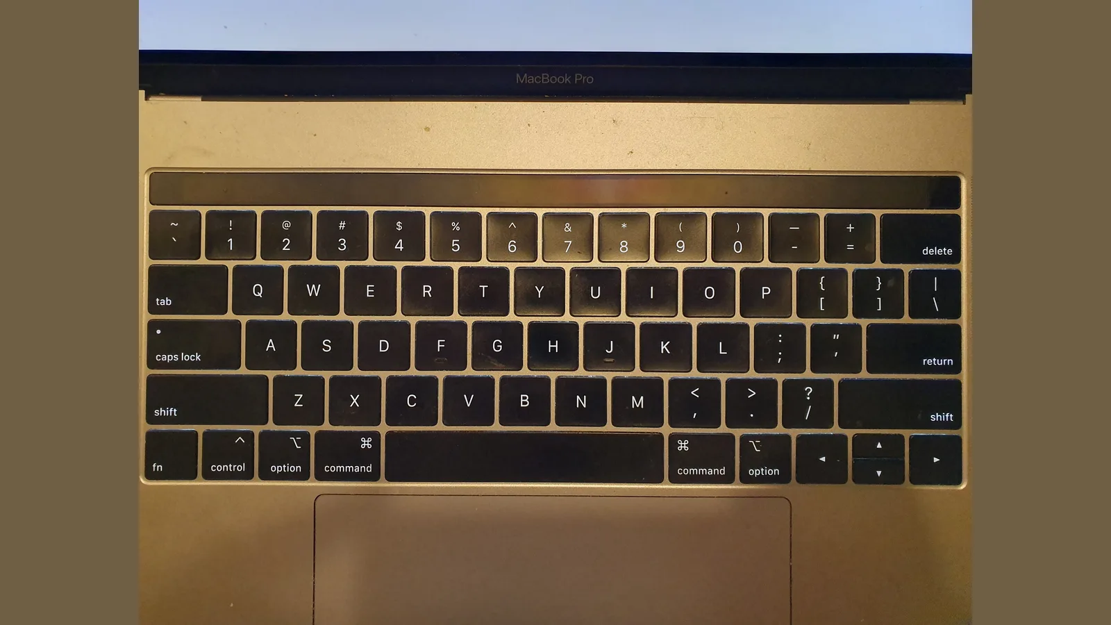 A MacBook Pro keyboard seen from directly above: black keys with some dust on them, a dark Touch Bar strip above the number keys, the top of the wide trackpad below, and above them the bottom edge of the open screen, with 'MacBook Pro' printed on its black border and a pale lit strip of screen at the top