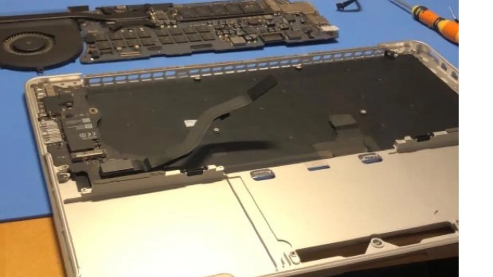 An aluminium laptop opened for repair on a blue mat: its logic board and cooling fan lifted out and lying above the empty case, a black flat cable curving across the inside, and an orange-handled screwdriver at the top right