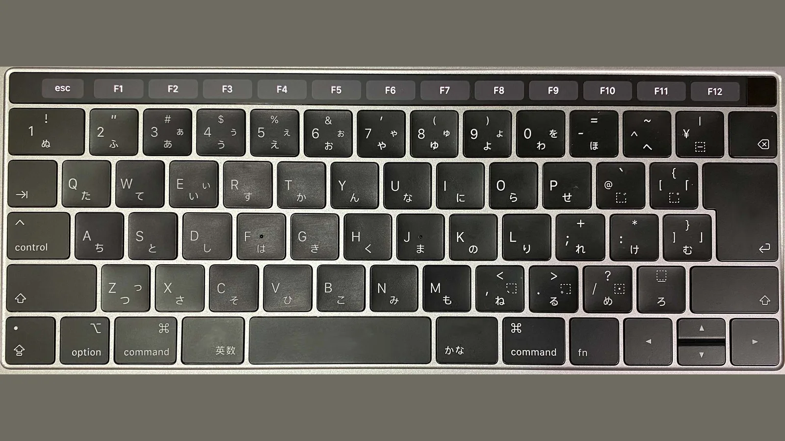 从正上方拍摄的 MacBook Pro 日文布局键盘。顶端本该是实体功能键的位置，是一条黑色的触控栏：左端显示 esc，右侧依次排着 F1 到 F12 的灰色按键标签