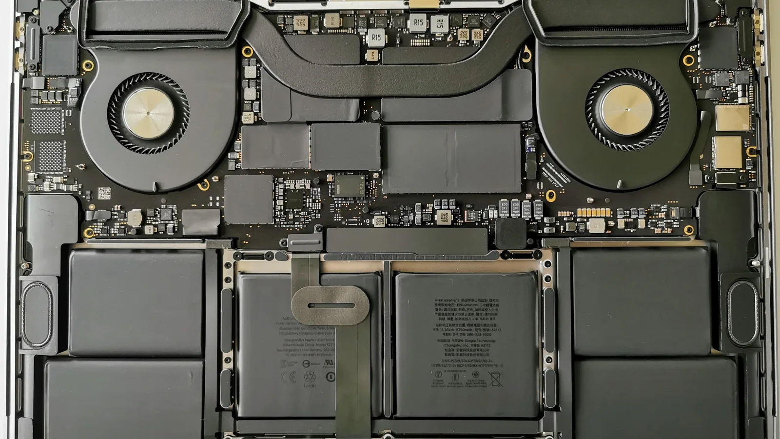 拆去底壳后从上方看到的 16 英寸 MacBook Pro 内部：上方两角各有一个圆形风扇，中间是主板与横跨其间的热管，下半部分排满六块扁平的电池单元