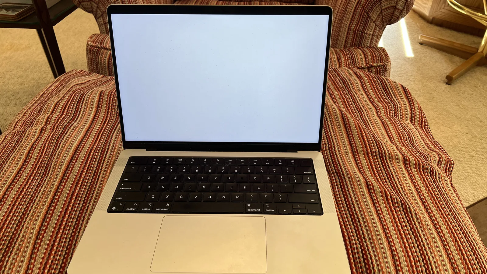 Sebuah komputer riba Apple berwarna perak yang terbuka di atas kerusi berlengan berjalur, skrinnya menyala putih kosong dari tepi ke tepi, dengan papan kekunci hitam dan trackpad yang lebar kelihatan