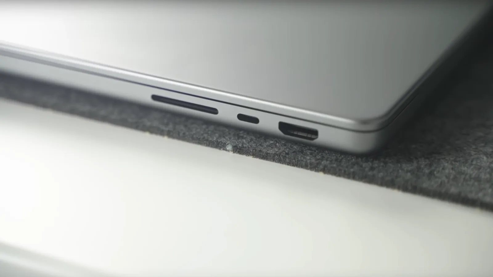 Gambar dekat tepi sebuah komputer riba Apple berwarna kelabu gelap yang tertutup di atas alas felt kelabu: slot kad, port USB-C dan port HDMI