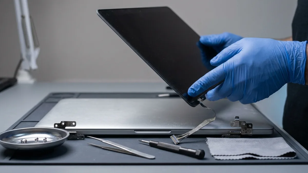 A blue-gloved hand lifting a laptop display panel clear of its base on a bench mat, its ribbon cable hanging free, with two hinges, tweezers, a screwdriver, a dish of screws and a cloth laid out below