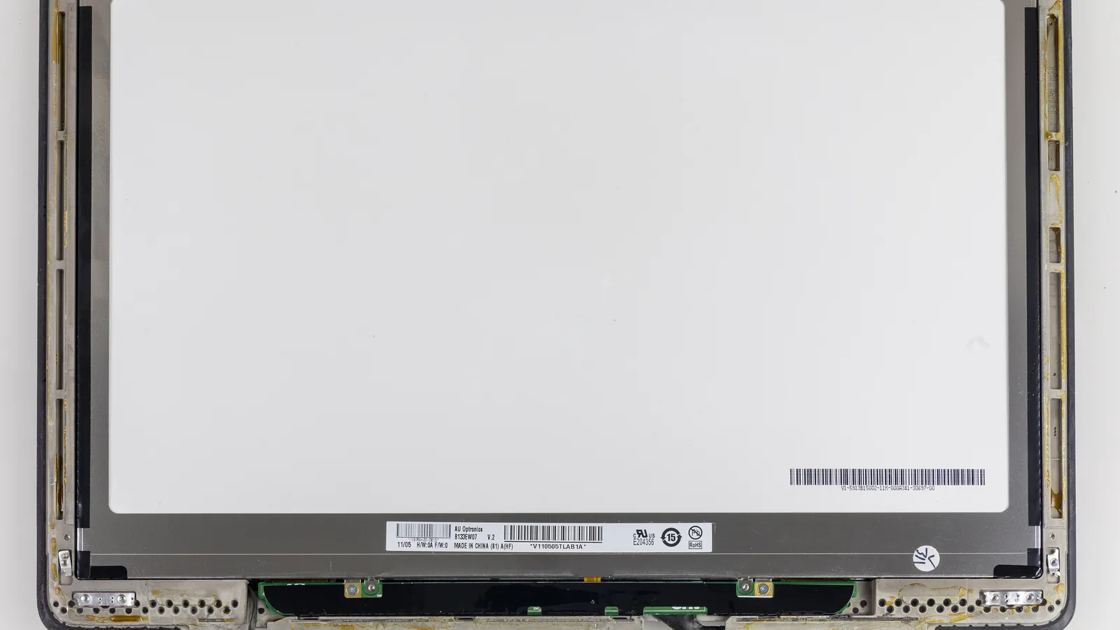A bare laptop LCD panel lying in its lid frame, seen from above: a large blank white sheet edged in grey metal, a white maker's label with barcodes near the bottom, a narrow green circuit board along the bottom edge and metal hinge brackets in the two lower corners