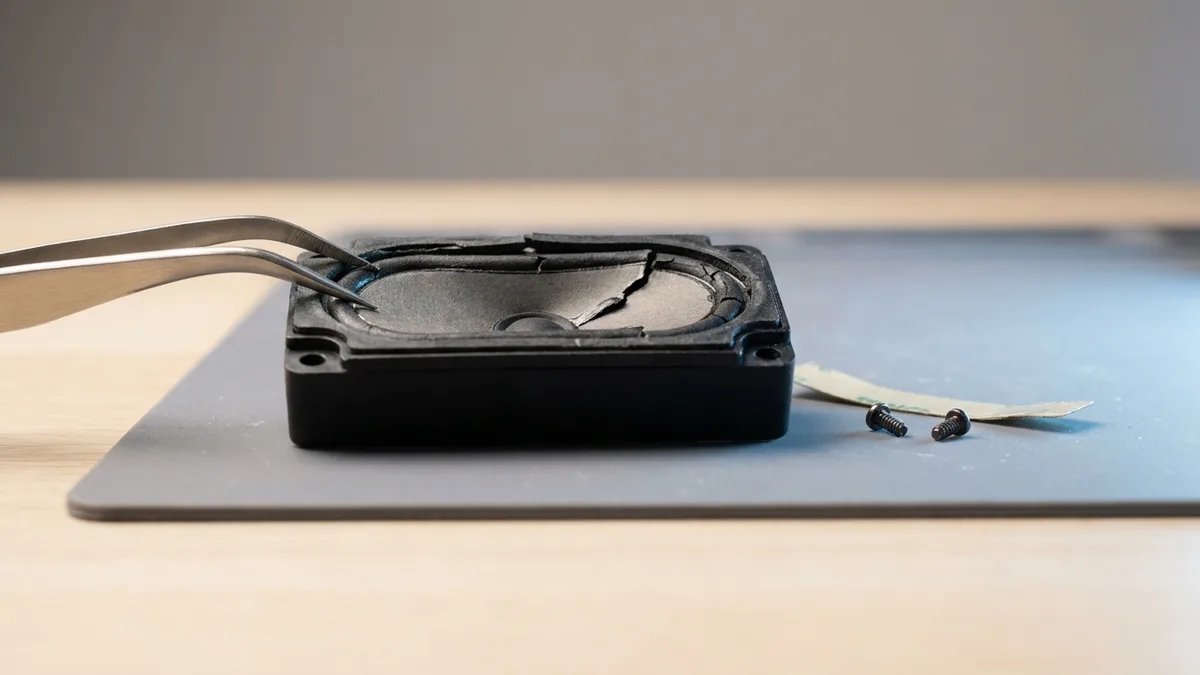 A laptop speaker module removed onto an anti-static mat, its diaphragm split and the rubber surround cracked