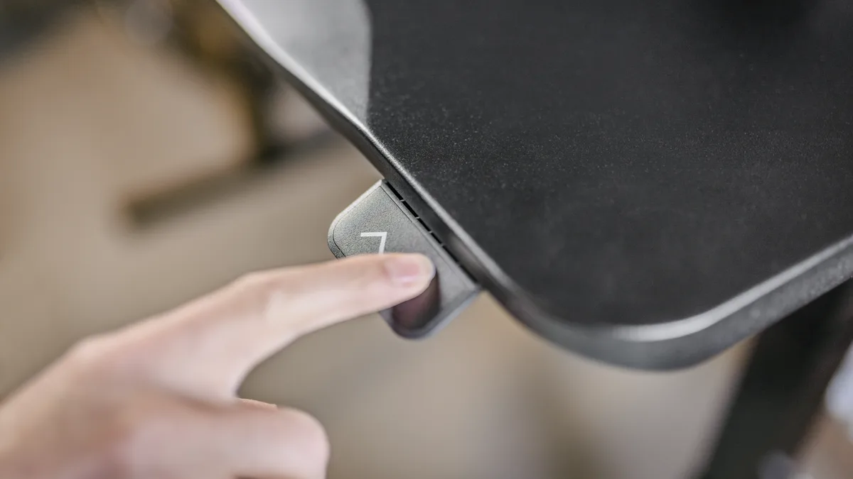 A finger pressing the height control recessed under the front edge of a black height-adjustable desktop