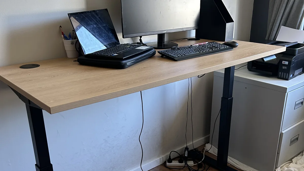 A height-adjustable desk raised to standing height with a laptop open flat on the desktop, a second monitor behind it and loose cables running down to a power strip on the floor