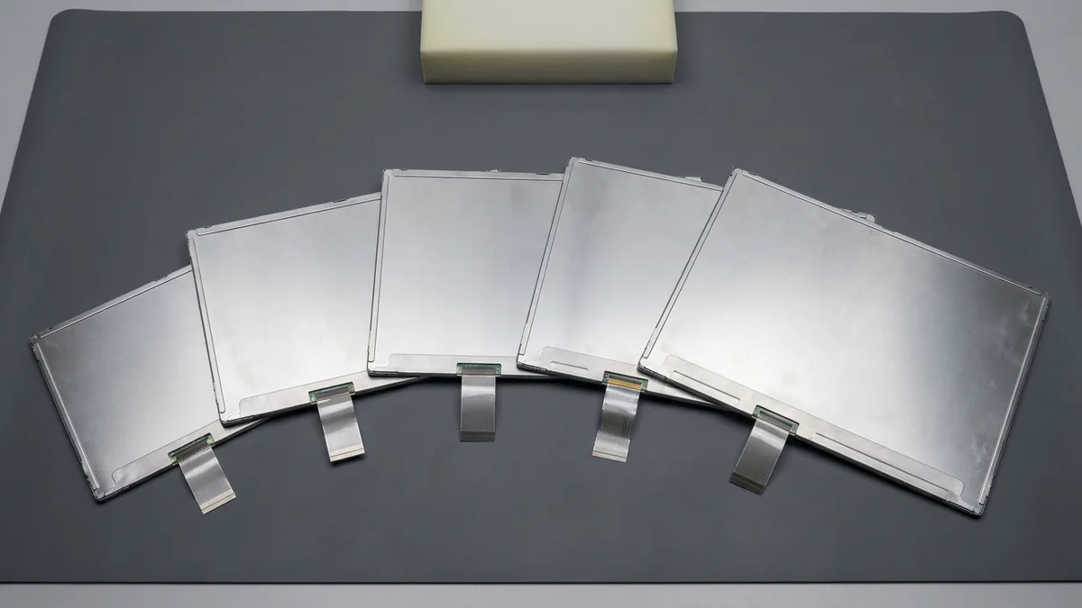 Five bare laptop display panels lie fanned out in a row on a dark grey bench mat, all face down so only their plain silver metal rear chassis show, each with a flat ribbon cable of a different width folded out along its lower edge, with a single foam block behind them.