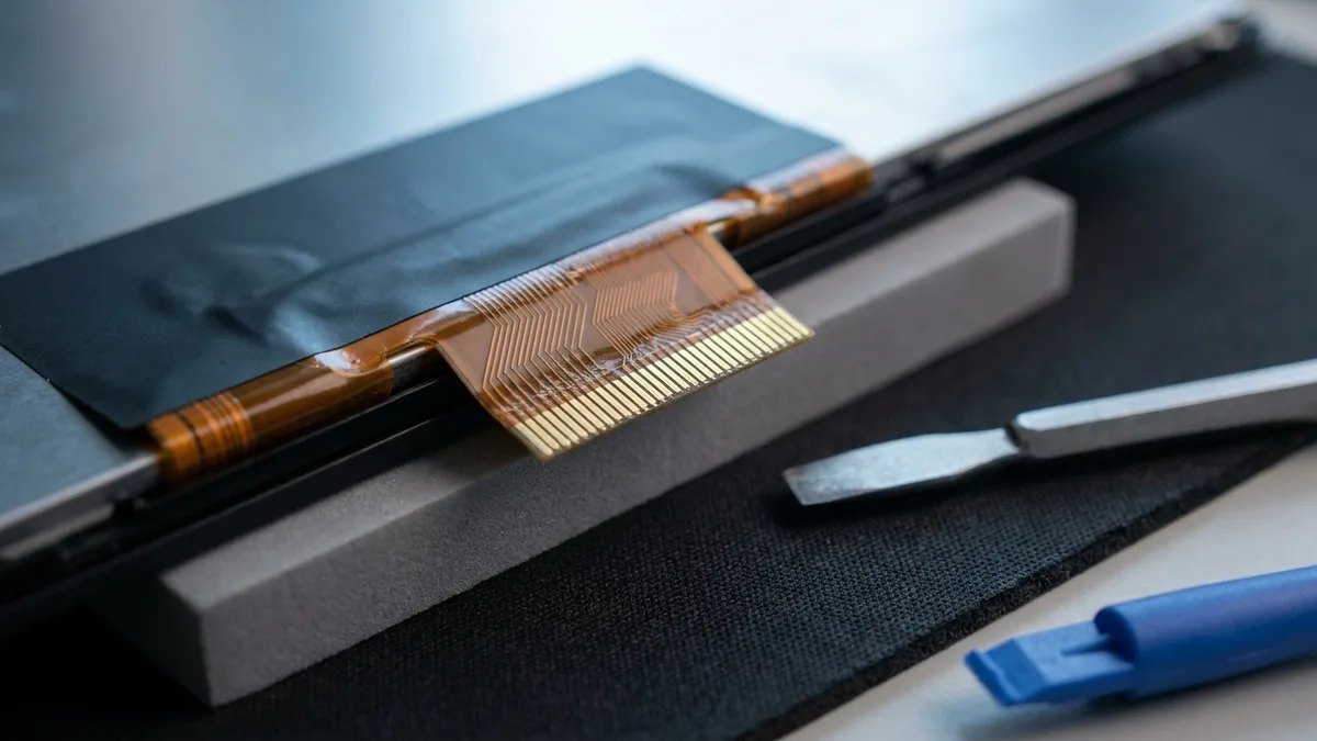 An extreme close-up of the lower edge of a bare laptop display panel lying face down, its amber flat ribbon cable lifted and folded back to expose a long row of gold connector pins and the fine copper traces inside the ribbon, with a plain metal spudger and a blue plastic pry tool on the black mat beside it.