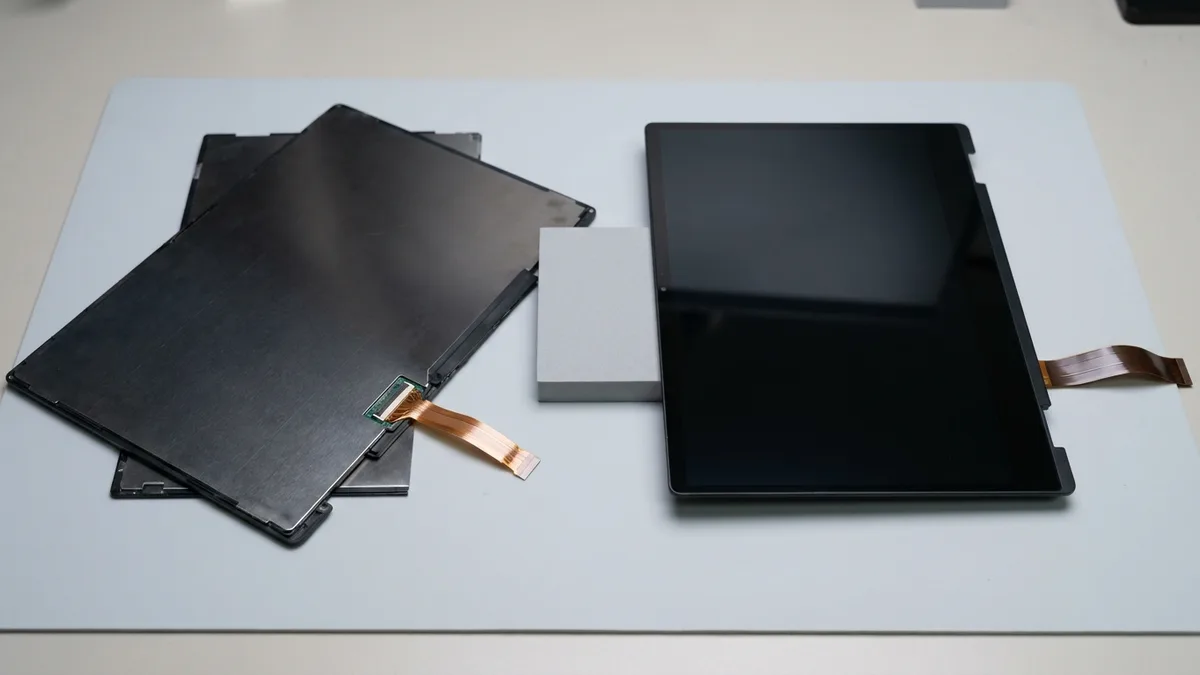 Two bare laptop display assemblies side by side on a pale bench mat: on the left a matte panel lying face down showing its bare dark rear and one copper ribbon; on the right a glossy touchscreen assembly face up but completely dark and switched off, with its own copper ribbon folded out from the edge, and a plain grey foam block between them.