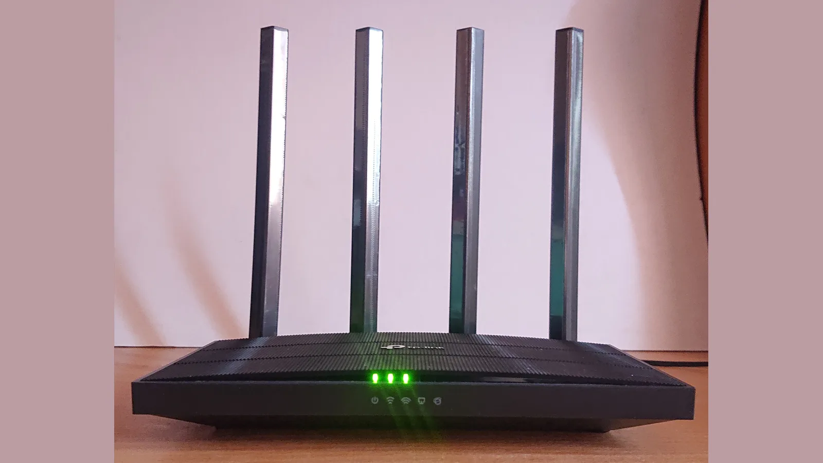 一台黑色 TP-Link 家用 Wi-Fi 路由器，竖着四根长天线，放在木桌上，背后是淡粉色的墙，正面亮着三盏绿色状态指示灯。