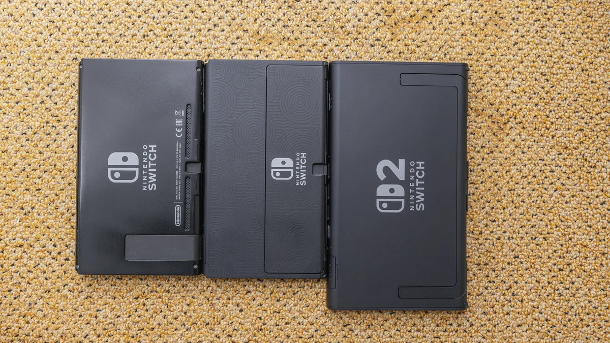 Three Nintendo consoles lying face-down side by side: the 2017 Switch on the left, the OLED model in the middle, and the visibly larger Switch 2 on the right