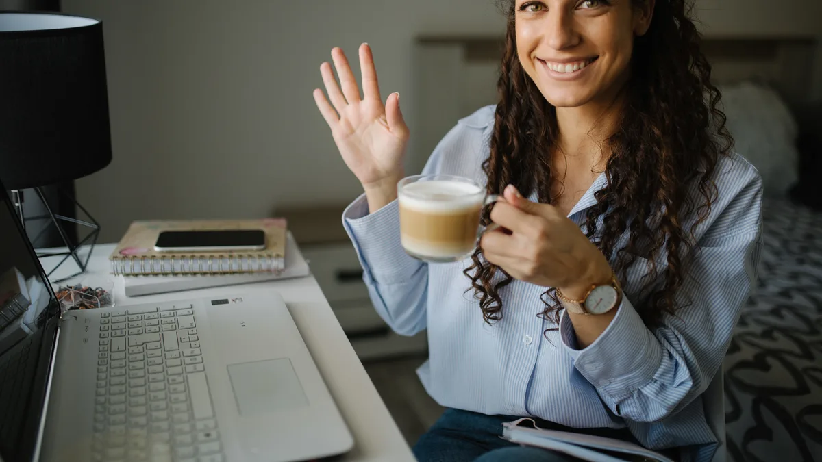 A woman at a home-office desk smiling with a cup of coffee, an open laptop, a notebook and a phone in front of her