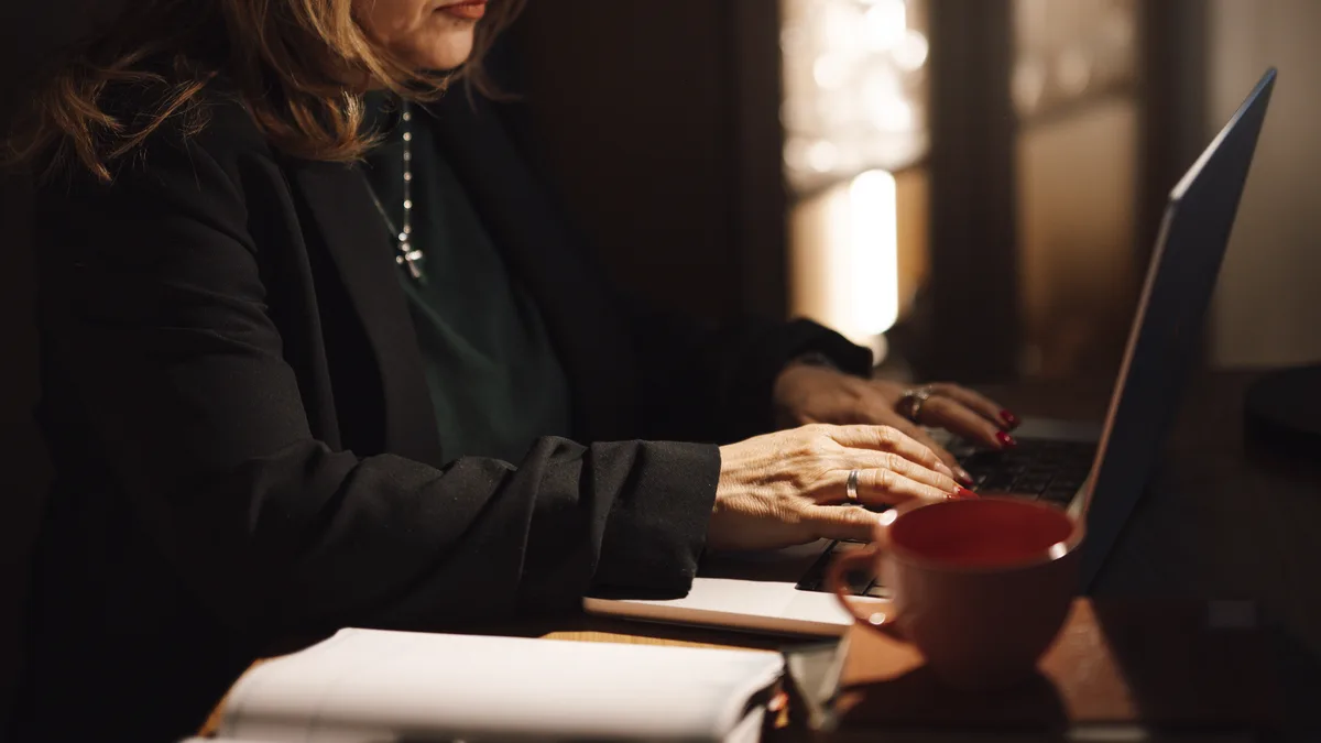A woman in business dress typing at a laptop beside papers and a mug, the screen angled away from the camera
